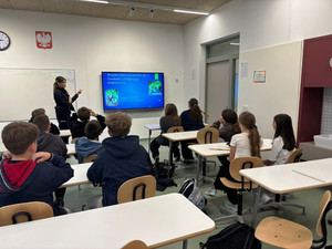 Zdjęcie przedstawia policjantkę stojącą przez uczniami siedzącymi w ławkach. W tle widać wyświetlony slajd prezentacji.