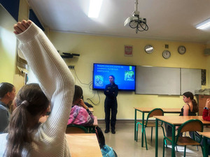 Zdjęcie przedstawia policjantkę stojącą przez uczniami siedzącymi w ławkach. W tle widać wyświetlony slajd prezentacji.