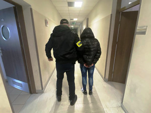 Zdjęcie przedstawia ubranego na ciemno policjanta, z opaską z napisem POLICJA na prawym ramieniu, który prowadzi przez korytarz, po swojej prawej stronie zatrzymaną kobietę. Kobieta ma kaptur założony a głowę. Oboje są tyłem do obiektywu aparatu.