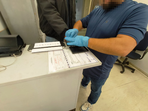 Zdjęcie przedstawia policjanta w ubraniu cywilnym, który wykonuje daktyloskopię u zatrzymanego mężczyzny.