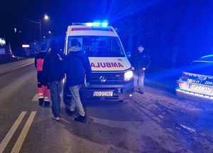 Zdjęcie przedstawia kilka osób, w tym umundurowanego policjanta stojącego przy radiowozie i karetce.