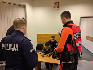 Zdjęcie przedstawia policjanta i ratownika medycznego stojących przy policjancie trzymającym na kolanach dziecko.