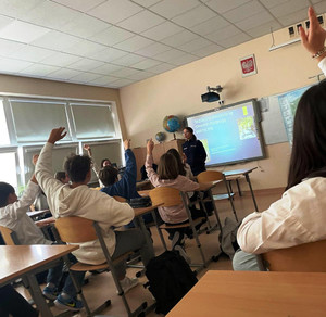 Zdjęcie przedstawia policjantkę stojącą przed uczniami siedzącymi w szkolnych ławkach. Część uczniów ma podniesione do góry ręce.