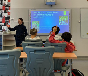 Zdjęcie przedstawia policjantkę stojącą przed uczniami siedzącymi w szkolnych ławkach. Jeden uczeń ma podniesioną do góry rękę.