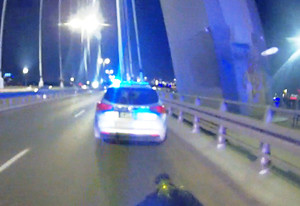 Zdjęcie przedstawia radiowóz z włączonymi światłami stojący na moście.