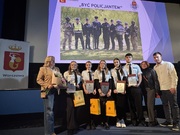 Zdjęcie przedstawia grupę osób, dorosłych oraz młodzieży ubranych w jasne koszule stojących ba scenie. Za nimi na slajdzie widnieje napis „Być Policjantem".