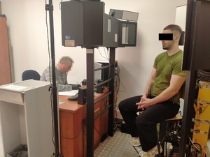 Zdjęcie przedstawia policjanta wykonującego daktyloskopię u zatrzymanego mężczyzny siedzącego na krześle.