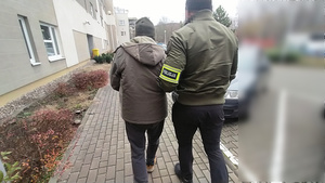 Zdjęcie przedstawia policjanta ubranego w cywilną odzież, z odblaskową opaską z napisem POLICJA na lewym przedramieniu, który prowadzi po swojej lewej stronie zatrzymanego mężczyznę.