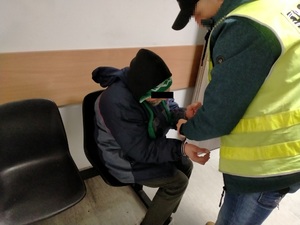 Zdjęcie przedstawia policjanta ubranego w odblaskową kamizelkę, który pochyla się nad siedzącym na krześle mężczyzną i zakłada mu kajdanki na rękach wyciągniętych do przodu.