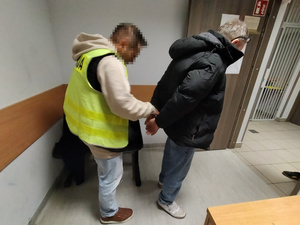 Zdjęcie przedstawia policjanta ubranego w odblaskową kamizelkę z napisem POLICJA, który zakłada kajdanki na ręce trzymane z tyłu zatrzymanemu mężczyźnie. Twarz policjanta jest zniekształcona komputerowym filtrem. Obaj znajdują się w pomieszczeniu dla osób zatrzymanych.