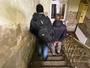 Zdjęcie przedstawia ubranego na ciemno policjanta, który sprowadza po schodach, idącego po jego prawej stronie zatrzymanego mężczyznę.