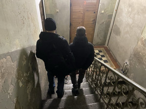 Zdjęcie przedstawia ubranego na ciemno policjanta, który sprowadza po schodach, idącego po jego prawej stronie zatrzymanego mężczyznę.