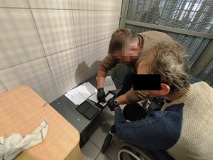 Zdjęcie przedstawia policjanta, który wykonuje odciski palców zatrzymanemu mężczyźnie. Mężczyzna ten ma część twarz zasłoniętą czarnym prostokątem.