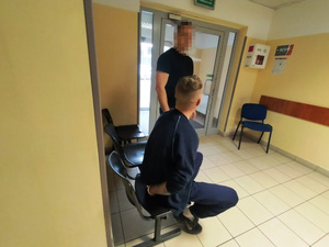 Zdjęcie przedstawia policjanta, który nadzoruje zachowanie zatrzymanego mężczyzny siedzącego na krześle w korytarzu przed pomieszczeniem dla osób zatrzymanych.