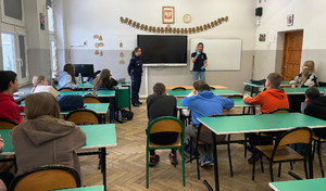 Zdjęcie przedstawia policjantkę i kobietę w niebieskich ochronnych rękawiczkach stojące przed uczniami siedzącymi w ławkach.
