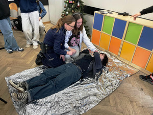 Zdjęcie przedstawia policjantkę i dziewczynkę klęczącą obok leżącej na podłodze uczennicy.