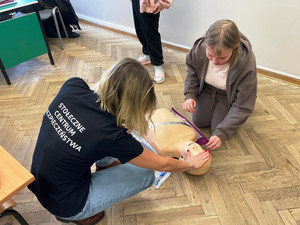 Zdjęcie przedstawia kobietę w koszulkę z napisem "stołeczne centrum bezpieczeństwa" i dziewczynkę klęczące obok leżącej na podłodze uczennicy.