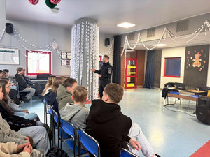 Zdjęcie przedstawia policjanta stojącego przed uczniami siedzącymi na krzesłach.