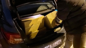 Zdjęcie przedstawia fragment sylwetki policjanta sięgającego do otwartego bagażnika, gdzie znajdują się kartonowe pudła.