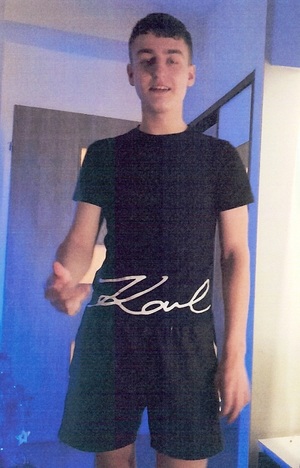 Zdjęcie przedstawia młodego chłopaka o ciemnych włosach, ubranego w ciemną koszulkę na krótki rękaw i krótkie ciemne spodenki. Chłopak znajduje się w pomieszczeniu.