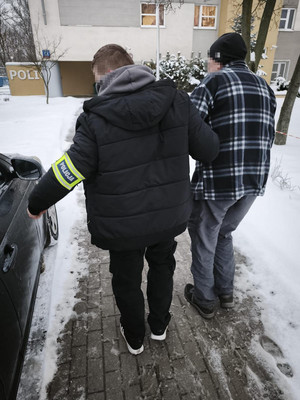 Zdjęcie przedstawia ubranego na ciemno mężczyznę z opaską POLICJA na lewym przedramieniu, który prowadzi po swojej prawej stronie starszego mężczyznę.