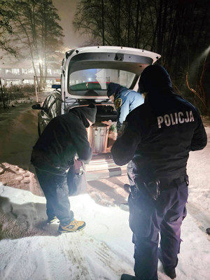 Zdjęcie przedstawia policjanta, mężczyznę oraz strażnika miejskiego stojących w otwartej klapie bagażnika samochodu. Na zewnątrz jest ciemno.