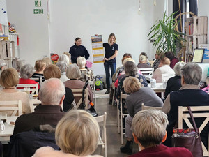 Zdjęcie przedstawia policjantkę i kobietę stojących przed siedzącymi na krzesłach seniorami.