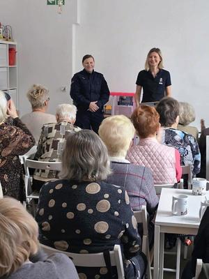 Zdjęcie przedstawia policjantkę i kobietę stojących przed siedzącymi na krzesłach seniorami.