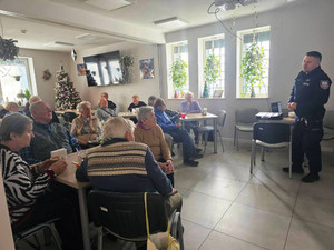 Zdjęcie przedstawia policjanta stojącego przed seniorami siedzącymi w fotelach na sali.