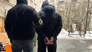 Zdjęcie przedstawia ubranego na ciemno policjanta, który prowadzi przez podwórze ubranego na ciemno zatrzymanego mężczyznę.