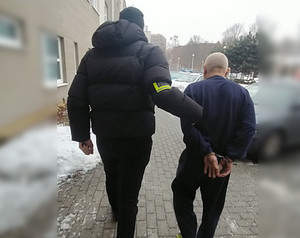 Zdjęcie przedstawia ubranego na ciemno policjanta, który prowadzi wzdłuż budynku ubranego w granatowy dres zatrzymanego mężczyznę.