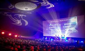 Zdjęcie przedstawia wielki ekran na gali, widać granatowe światła, które oświetlają salę.