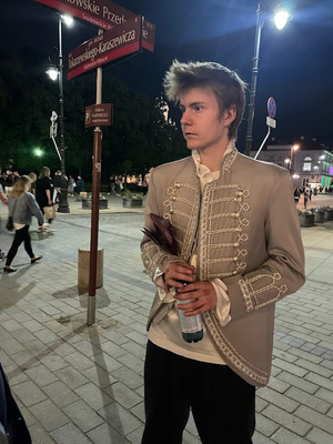 Zdjęcie przedstawia ubranego w beżowy blezer chłopaka. W tle widać słup z tabliczkami przedstawiającymi skrzyżowanie ulic.