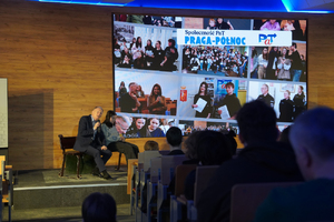 Zdjęcie przedstawia mężczyznę z mikrofonem przy ustach, siedzącego na ławce obok młodej dziewczyny. Tło, za ich plecami stanowi kolorowa grafika.