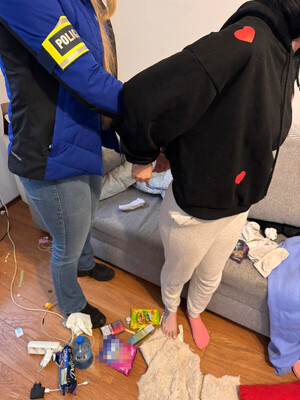 Zdjęcie przedstawia policjantkę, z opaską POLICJA na przedramieniu, która zakłada kajdanki kobiecie stojącej do niego tyłem.