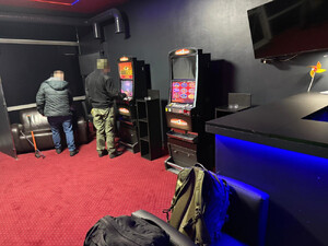 Zdjęcie przedstawia dwie osoby stojący przy automatach do gier hazardowych, umieszczonych w pomieszczeniu przy ścianie.