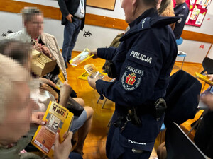 Zdjęcie przedstawia policjantkę, która wręcza siedzącej na krześle kobiecie ulotkę.