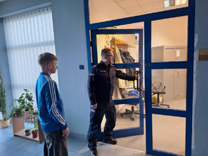 Zdjęcie przedstawia policjanta, który uchyla przeszklone drzwi, niedaleko niego stoi chłopak ubrany w niebieską bluzę.