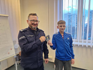 Zdjęcie przedstawia policjanta i nastolatka, którzy wspólnie trzymają policyjny emblemat.