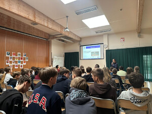 Zdjęcie przedstawia policjantkę z mikrofonem, stojącą przed siedzącymi na krzesłach uczniami.