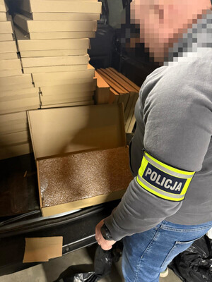 Zdjęcie przedstawia kartony ułożone w bagażniku samochodu i stojącego obok samochodu mężczyznę z opaską POLICJA na lewym przedramieniu.