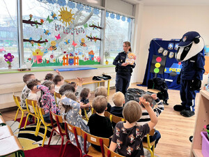 Zdjęcie przedstawia policjantkę i misia stojących przed dziećmi siedzącymi na dywanie.