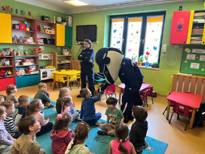 Zdjęcie przedstawia policjantkę i misia stojących przed dziećmi siedzącymi na dywanie.