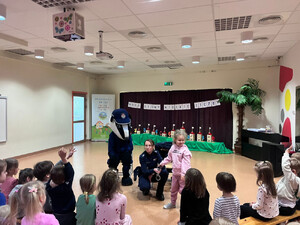 Zdjęcie przedstawia policjantkę i misia stojących przed dziećmi siedzącymi na dywanie.