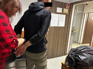 Zdjęcie przedstawia policjantkę ubraną w odzież cywilną, która zakłada kajdanki mężczyźnie stojącemu do niej tyłem. Część twarzy mężczyzny zasłania czarny prostokąt.