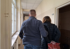 Na zdjęciu widać policjanta w ubraniu cywilnym, który prowadzi po swojej prawej stronie, przez korytarz zatrzymaną kobietę.