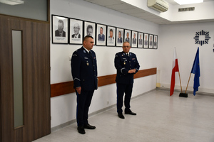 Zdjęcie przedstawia dwoje umundurowanych policjantów stojących na tle ściany, na której widnieją portrety policjantów.