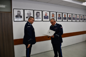 Zdjęcie przedstawia dwoje umundurowanych policjantów, podających sobie dłonie i stojących na tle ściany, na której widnieją portrety policjantów.