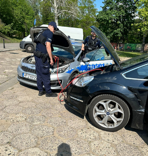 Zdjęcie przedstawia dwóch policjantów, którzy pochylają się nad otwartą maską radiowozu.