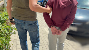 Zdjęcie prowadzi policjanta prowadzącego po swojej prawej stronie zatrzymanego mężczyznę. Mężczyzna ten ma kajdanki na rękach trzymanych za plecami.